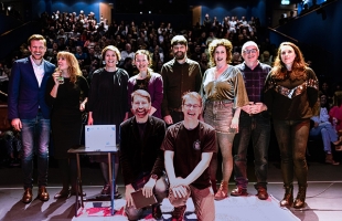 A group of smiling people in two rows on the stage at Pavilion Theatre, with an out-of-focus, packed auditorium behind them. It's the storytellers of the first Dublin Story Slam at Pavilion Theatre in February 2024, photographed at the conclusion of the evening.