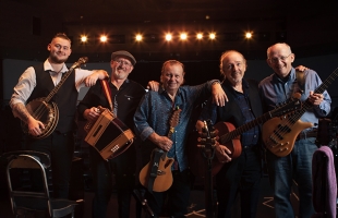 A photo of the five members of The Fureys, each holding their respective instrument (banjo, accordion, two with acoustic guitar, and bass guitar), smiling and leaning on one another, in what appears to be a music venue or theatre.