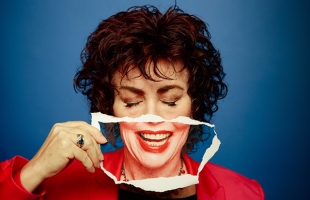 Ruby Wax in a bright red jacket, holding a piece of paper with a toothy grin on it in front of her own mouth.