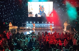 ABBA Forever performing for a packed audience in white outfits, with a projector screen behind them.