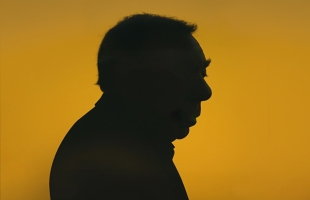 A side-profile silhouette of Andrew Lloyd Webber looking down against a bright, warm amber-yellow background. The lighting creates a dramatic, moody contrast, casting his head and shoulders entirely in shadow.