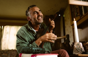 Aindrias de Staic, a smiling man with greying hair, sits in a warmly lit room, holding and playing a violin. A lit candle stands on the table beside him along with stacked books and a small record player, giving the cosy interior a rustic, intimate atmosphere. He looks upward as he plays, appearing relaxed and joyful.