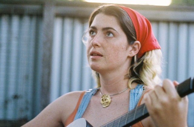 Roisín Gowen photographed from the chest up holding an acoustic guitar, viewed from a slight angle. The person wears a sleeveless top and a red headscarf, with a round pendant necklace visible at the neckline. One hand grips the guitar neck in the foreground. The background shows a corrugated metal fence or wall, softly out of focus, with warm natural light illuminating the scene.