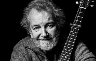 A black-and-white portrait of Andy Irvine who has a beard and tousled hair, smiling gently while holding a guitar close to his face.