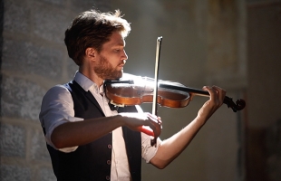 Anton Jablokob playing a violin under warm, dramatic lighting indoors, wearing a white shirt and dark vest while focusing on the performance