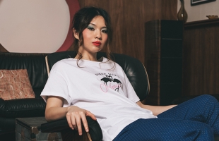 Belle Chen sits relaxed in a black leather chair in a cozy, mid-century styled room. She wears a white graphic T-shirt and blue patterned pants, with her hair tied back and bold makeup. One arm rests on the chair’s armrest while she gazes confidently toward the camera.
