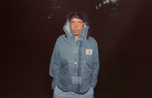 Bonnie Prince Billy Stands centre of a film image, he is wearing a long denim coat with the hood up, he is staring towards the camera intently with his hands in his pockets
