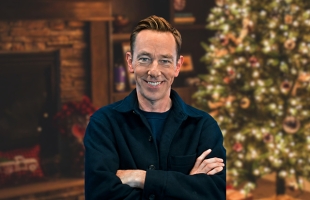 Ryan Tubridy smiles at the camera with his arms crossed in a medium shot. He wears a dark navy button-up jacket over a dark top. The background features a cozy, softly blurred indoor holiday scene, including a stone fireplace on the left and a decorated Christmas tree with lights and ornaments on the right.