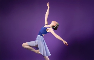 A dancer, in shades of blue and purple, with a purple background, strikes a pose. With right arm raised, and right foot pointed.