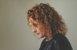 Briana Corrigan, a women with shoulder-length curly red hair shown in side profile, looking downward. She is wearing a dark jacket against a plain, light-coloured background, with soft, natural lighting creating a calm, reflective mood.