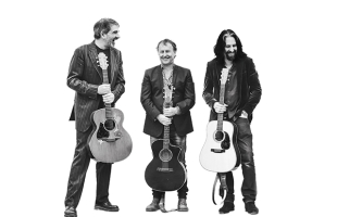 From left to right, Fiachna, Tom and Alan are standing together dressed in suit jackets holding their guitars, they are all smiling. The photo is black and white against a brilliant white background.