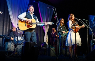 Cash Returns tribute band performing live on stage. JP Mac, portraying Johnny Cash, stands at the front left playing an acoustic guitar and singing, while Karen Martin, as June Carter Cash, stands to the right in a white dress playing a mandolin. A full band including drummer, bassist, and backing vocalist perform behind them, with colourful stage lighting and draped curtains enhancing the scene.
