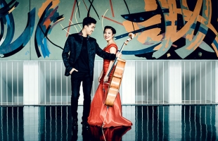 Bryan Cheng, wearing an all black suit and holding his cello out beside him, stands alongside Silvie Cheng dressed in a long red silk gown
