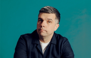 A studio portrait of a Chris Kent, a man with short dark hair and light stubble, wearing a dark jacket over a light shirt. He faces slightly to one side with a neutral expression. The background is teal with soft, even lighting.