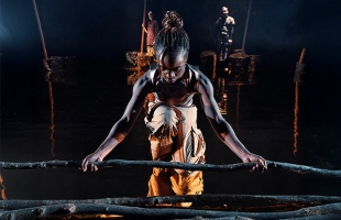 A young black female dancer, dressed in a gold top, and orange trousers bends down to pick up a large stick