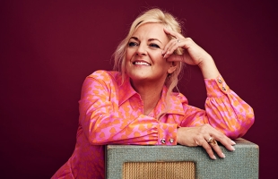 Claire Martin, A smiling blonde woman in a bright pink patterned blouse, leans on a vintage-style speaker against a deep red background.