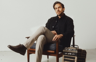 Man with dark hair and light stubble sits relaxed in a wooden armchair against a plain light background. He wears a black jacket, dark shirt, beige trousers, and brown leather shoes, with one leg crossed over the other. His expression is calm and thoughtful. A bandoneon (accordion-like instrument) rests on the floor beside him.