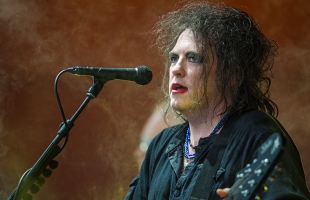 Robert Smith of The Cure performs onstage, singing into a microphone while playing guitar. He has tousled dark hair, smudged eye makeup and red lipstick, and wears a loose black shirt with layered blue necklaces. Warm stage lighting and haze create a moody concert atmosphere.