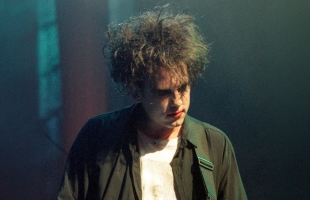 Robert Smith with tousled dark hair and pale makeup performing on stage, wearing a black shirt over a white top, looking downward under moody blue-green concert lighting.