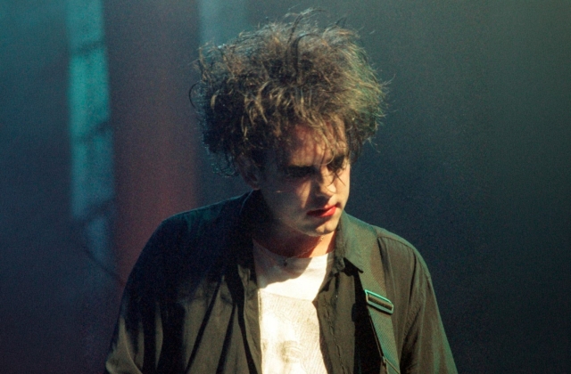 Robert Smith with tousled dark hair and pale makeup performing on stage, wearing a black shirt over a white top, looking downward under moody blue-green concert lighting.