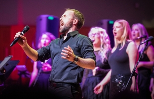 A bearded male vocalist in a black shirt sings passionately into a handheld microphone with his eyes closed and free hand raised. Behind him on stage, several female backing vocalists in black outfits sing along under vibrant purple and red stage lighting.