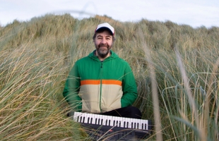 David O'Doherty is sat on a sand dune, surrounded by long grass, and his keyboard on the his lap