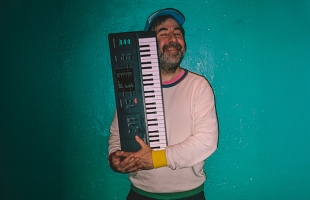 David O'Doherty stands wearing a cap & jumper. He is hugging his keyboard with a happy smile leaning against a green coloured wall.