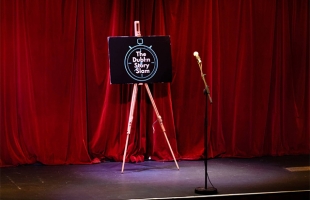 The image shows a theatre stage with red velvet curtain across the back. On stage there is a microphone and an easel with an image that reads ' Dublin story slam'.