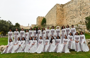 Two lines of women, dressed in traditional outfits pose with a stone walled structure behind them