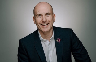 A portrait of David, he sits wearing a suit, he is smiling towards the camera