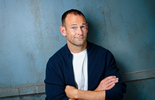 David Nihill with his arms crossed, smirking at the camera, with a blue wall behind him