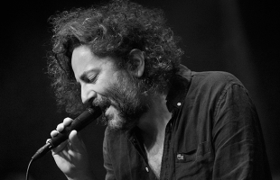 Black-and-white photograph of a person performing on stage, shown in close-up profile while holding a handheld microphone near the mouth. The person has curly hair and facial hair and wears a dark, buttoned shirt with an open collar. The microphone cable runs downward out of frame. Soft stage lighting illuminates the face and upper body, while the background fades into deep shadow, emphasizing the performer and the microphone in the foreground.