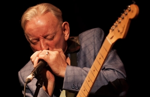 Don Baker playing the harmonica, with a guitar hanging in front of him