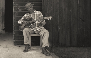 A black and white image of Eric Bibb sitting with his guitar wearing a checked shirt, in a typical