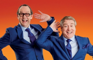 Eric & Ern in matching blue suits pose playfully with raised hands against a bright orange gradient background.