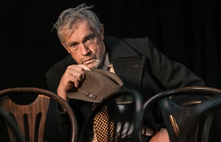Seamus O'Rourke, dressed in a dishevelled brown suit, sits on a wooden chair, surrounded by empty chairs, looking despondent. He is holding his hat in his hand.