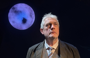 Seamus O'Rourke, dressed in a grey suit, stands in front of a blue full moon