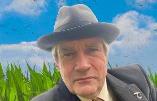 Seamus O'Rourke, dressed in a grey trilby hat, and suit, with a small lapel pin, with a cloudy blue sky and green field behind him