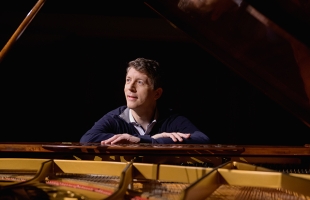 Finghin Collins sits at an open grand piano, casually dressed and looking off the left, with his arms resting on the top