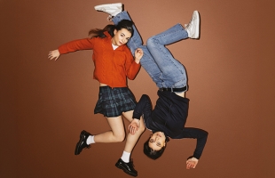 Two young women are captured mid-air against a plain brown background, creating a playful, gravity-defying effect. One, in a red cardigan and plaid skirt, appears upright, while the other, in a dark top and jeans, is upside down.