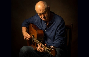 A middle-aged or older man with a shaved head, wearing a dark blue button-down shirt, sitting and playing an acoustic guitar against a dark background. He is looking down at his hands as he frets the strings.
