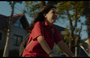 A young girl, wearing a red polo shirt, and a brown backpack, ride a bike through a suburban street, wind blowing in her hair