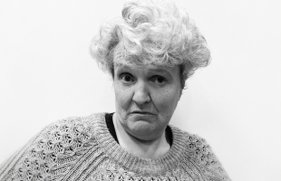 Rose Henderson wearing a knitted jumper, staring quizzically down the camera lens. The image is in black and white.