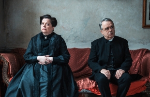 A sternly dressed woman and a clergyman sit side by side on a red sofa, their distant gazes and rigid postures suggesting unease or emotional separation.