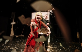 Two actors from Hay! stand on stage looking towards the camera, the woman wears a headband and a red dress, she is holding a basket full of confettis and its throwing it towards the camera, she is smiling. The man stands behind her in a suit catching the confetti in the air