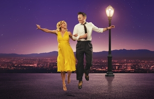 Liza Pullman, dressed in a yellow dress, and Joe Stillgoe, in a suit, arm in arm, dancing by a streetlight