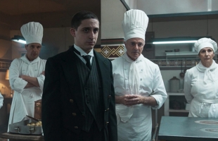 A man in a formal black suit, waistcoat, and white-collared shirt stands prominently in the foreground of a professional kitchen with a serious expression. Surrounding him are three kitchen staff members dressed in traditional white chef jackets and tall white toque hats: a man with arms crossed to the left, a man standing central with hands clasped in front of him, and a woman standing to the right.