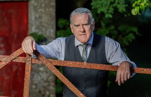 Mikel Murfi stands with his hands rested over a rusted gate, in a rural garden, wearing a suit