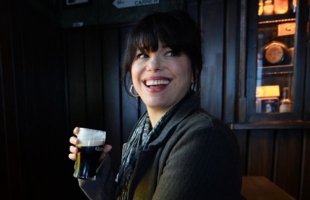 A smiling woman with dark hair and bangs sitting inside a pub, holding a small glass of Guinness stout. She is wearing a jacket, a patterned scarf, and hoop earrings, looking off to the side with a broad smile. Dark wooden paneling and a display cabinet are visible in the background.