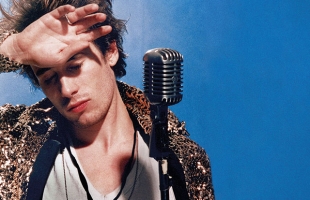 A close up of Jeff Buckley, he stands in front of a microphone with a his hand across his forehead and his eyes closed looking off to the distance. He's wearing a white t-shirt, snake skin pattern jacket against a blue background.